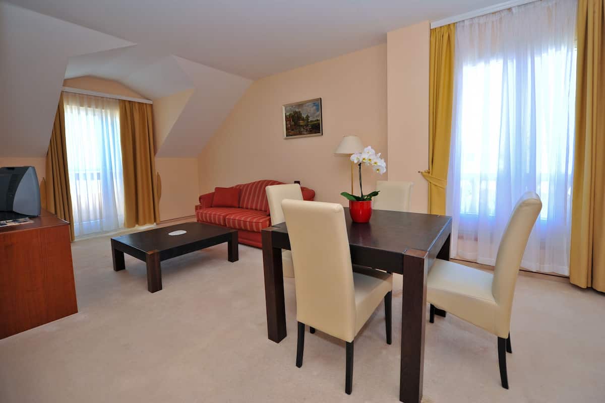 A hotel room with a dining table, two chairs, a sofa, a TV, and a lamp.