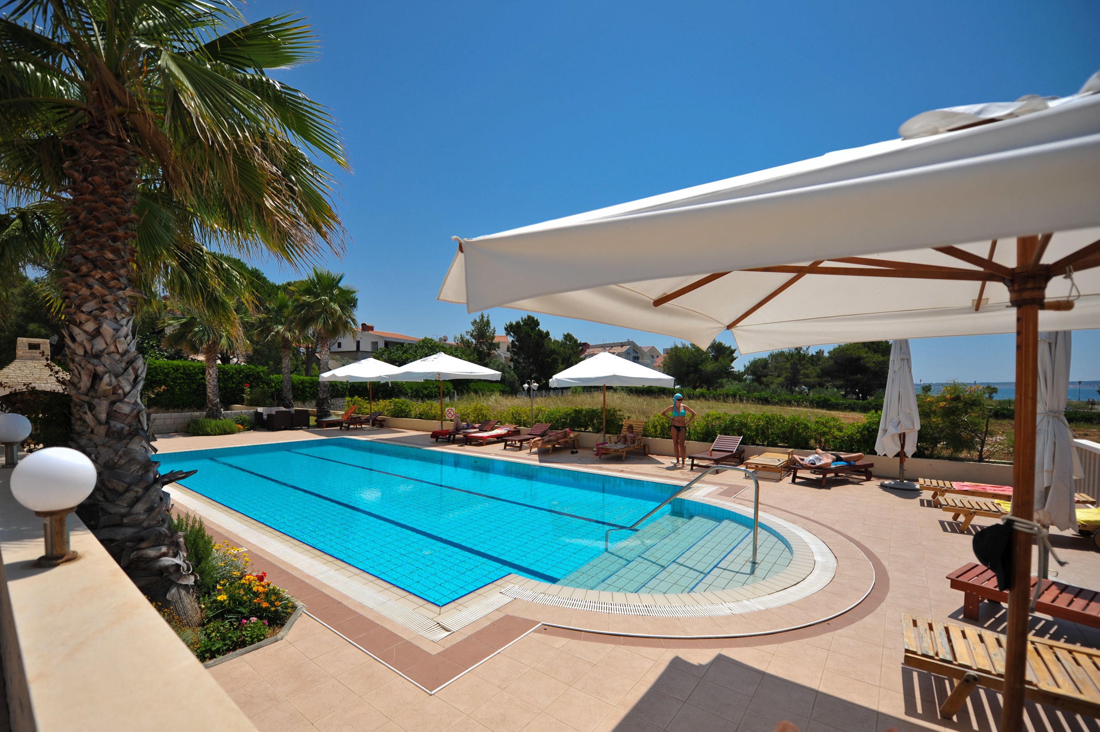 A swimming pool with sun loungers and umbrellas, surrounded by palm trees and a clear sky.
