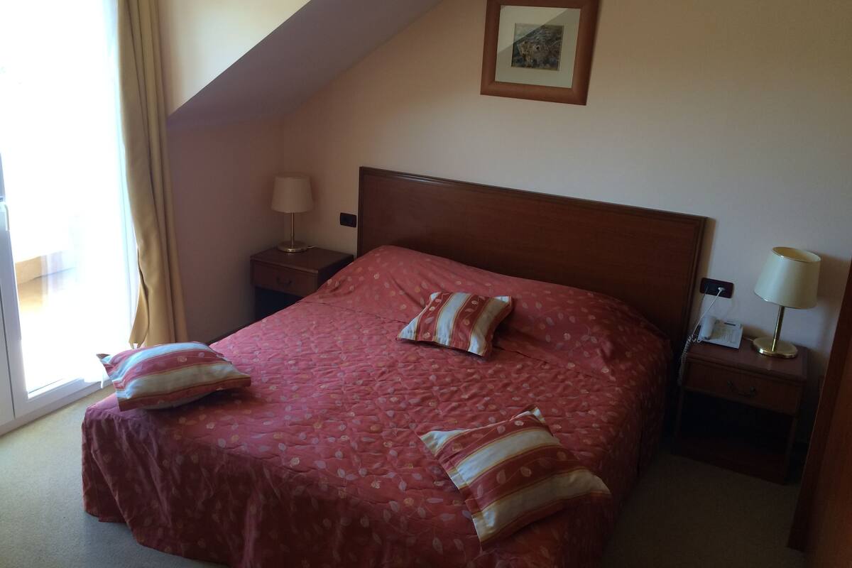 A hotel room with a bed, two bedside tables, a lamp on each table, a window with curtains, and a framed picture on the wall.