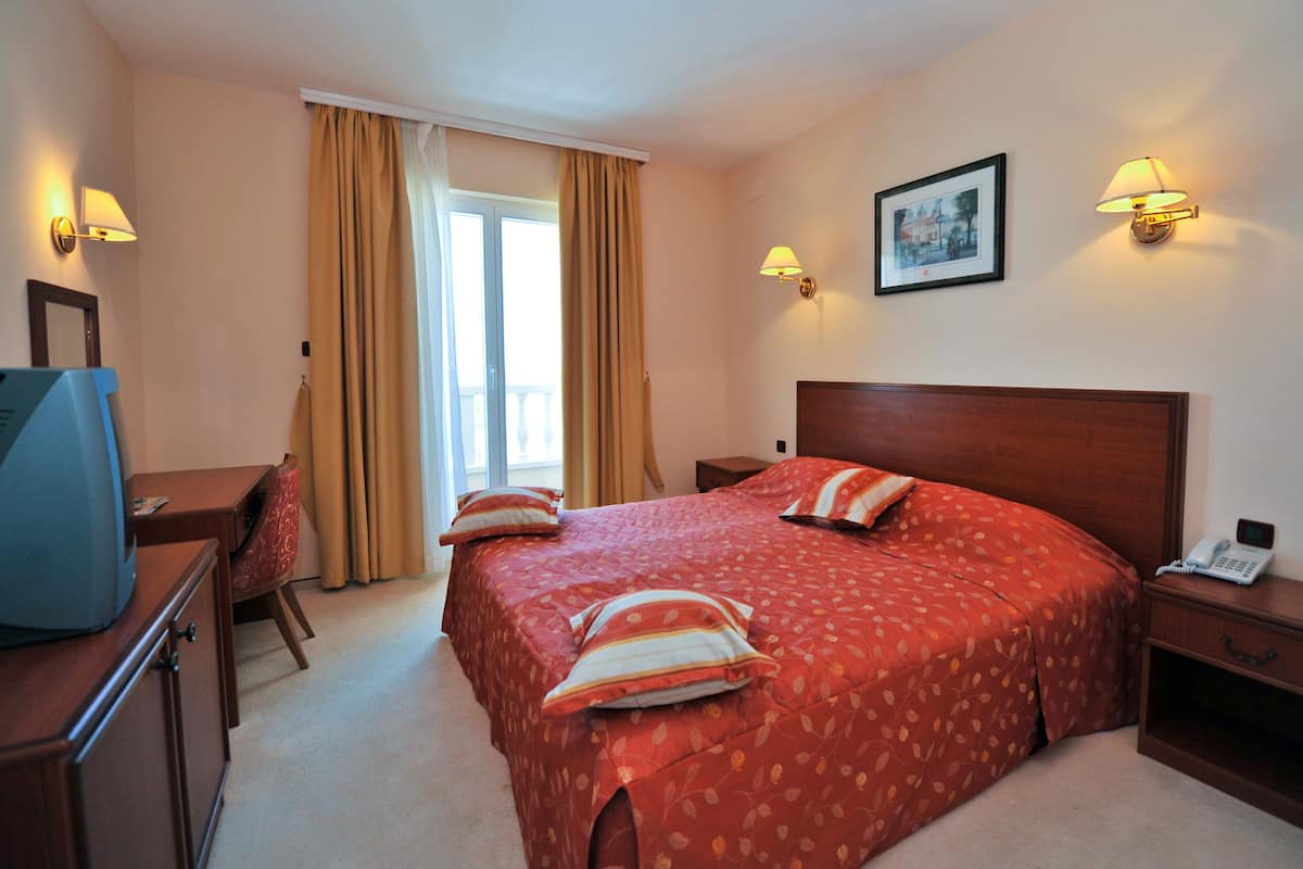 A hotel room with a large bed, two bedside lamps, a desk with a television, a chair, and a window with curtains.
