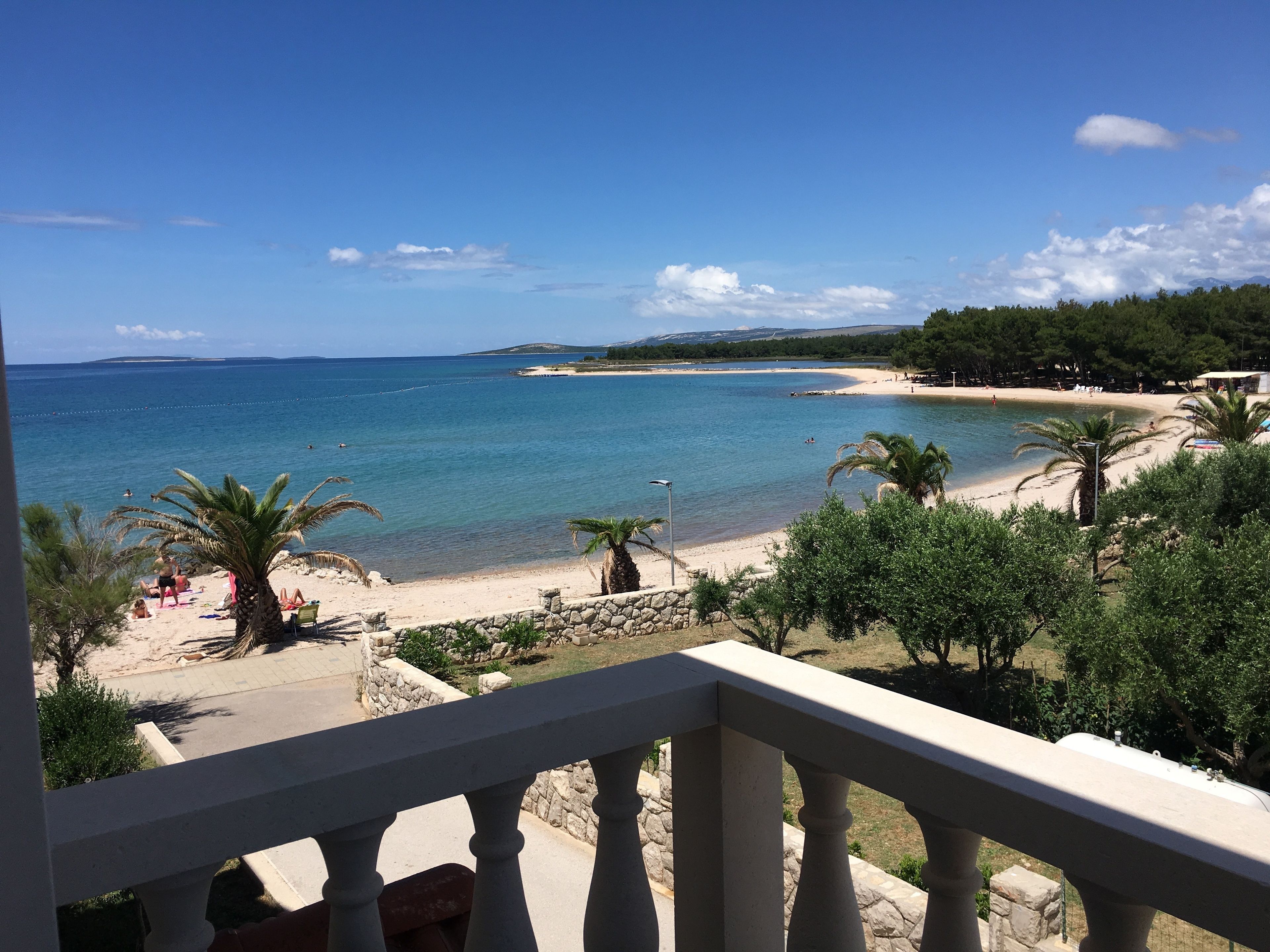 A beach view from a balcony with a stone balustrade, clear blue sea, sandy beach, and palm trees.