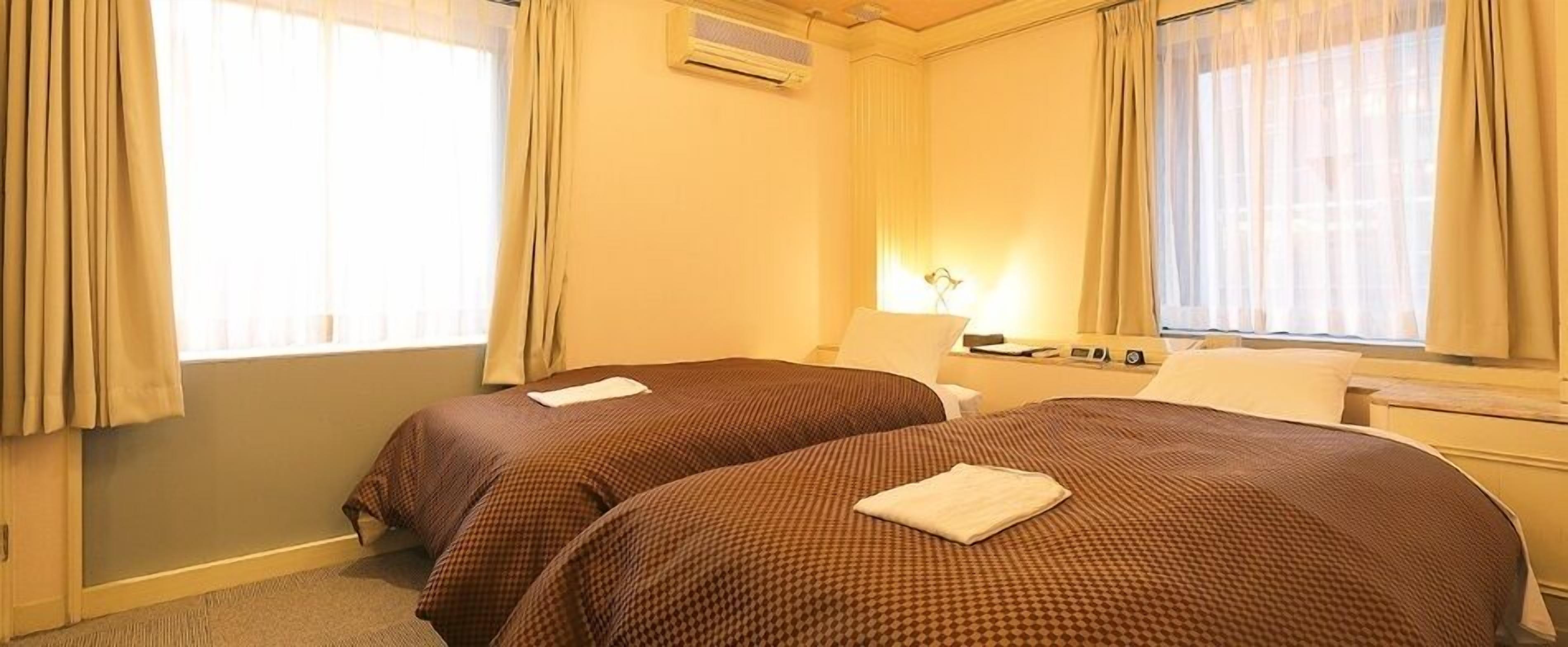 Camera d'albergo con due letti, una finestra con tende, un condizionatore d'aria e un comodino con una lampada.