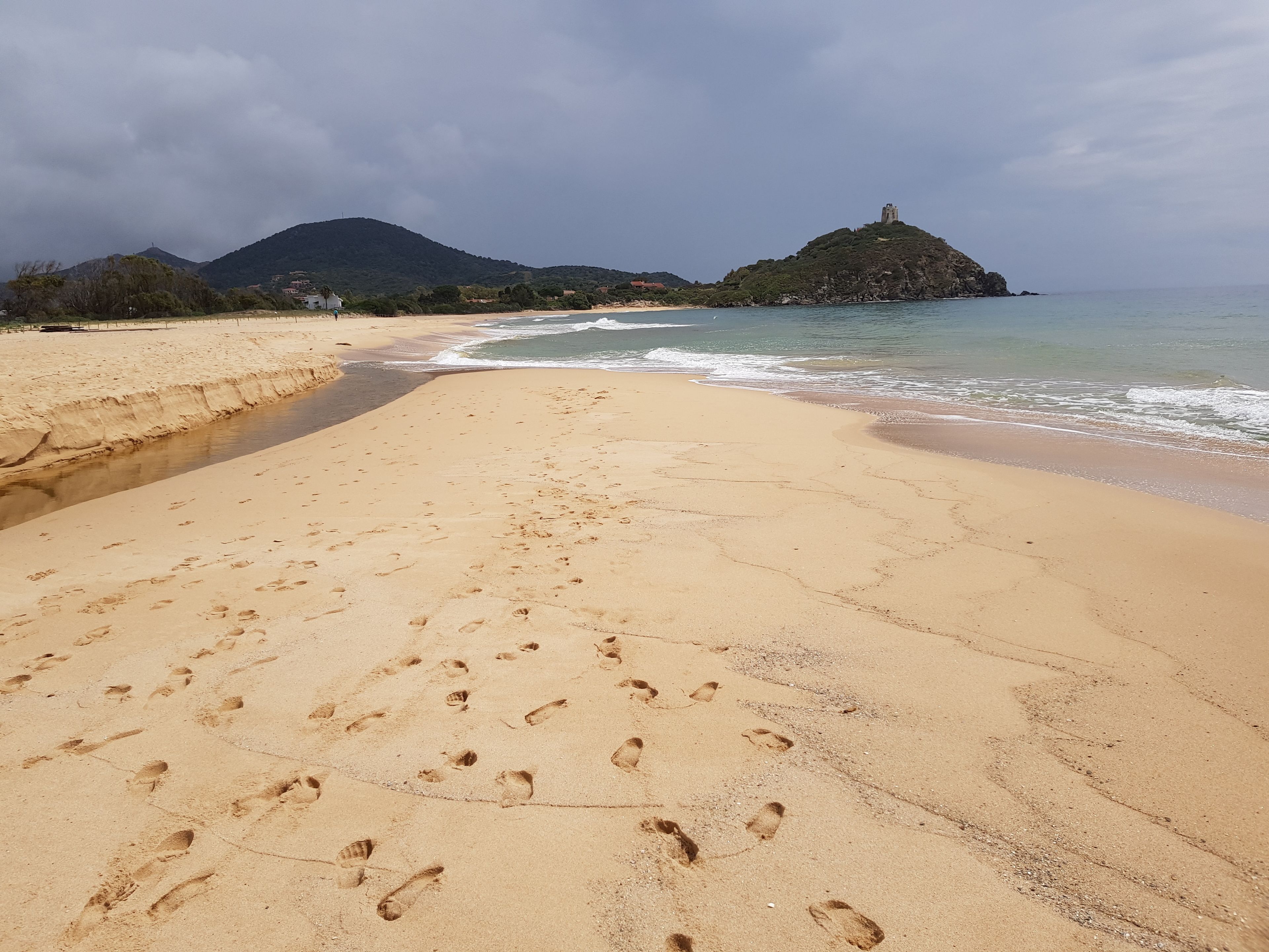 A sandy beach with footprints leading to the water, a rocky island with a tower in the distance, and a cloudy sky.