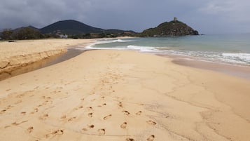 Una playa arenosa con huellas que se dirigen al agua, una isla rocosa con una torre a lo lejos y un cielo nublado.