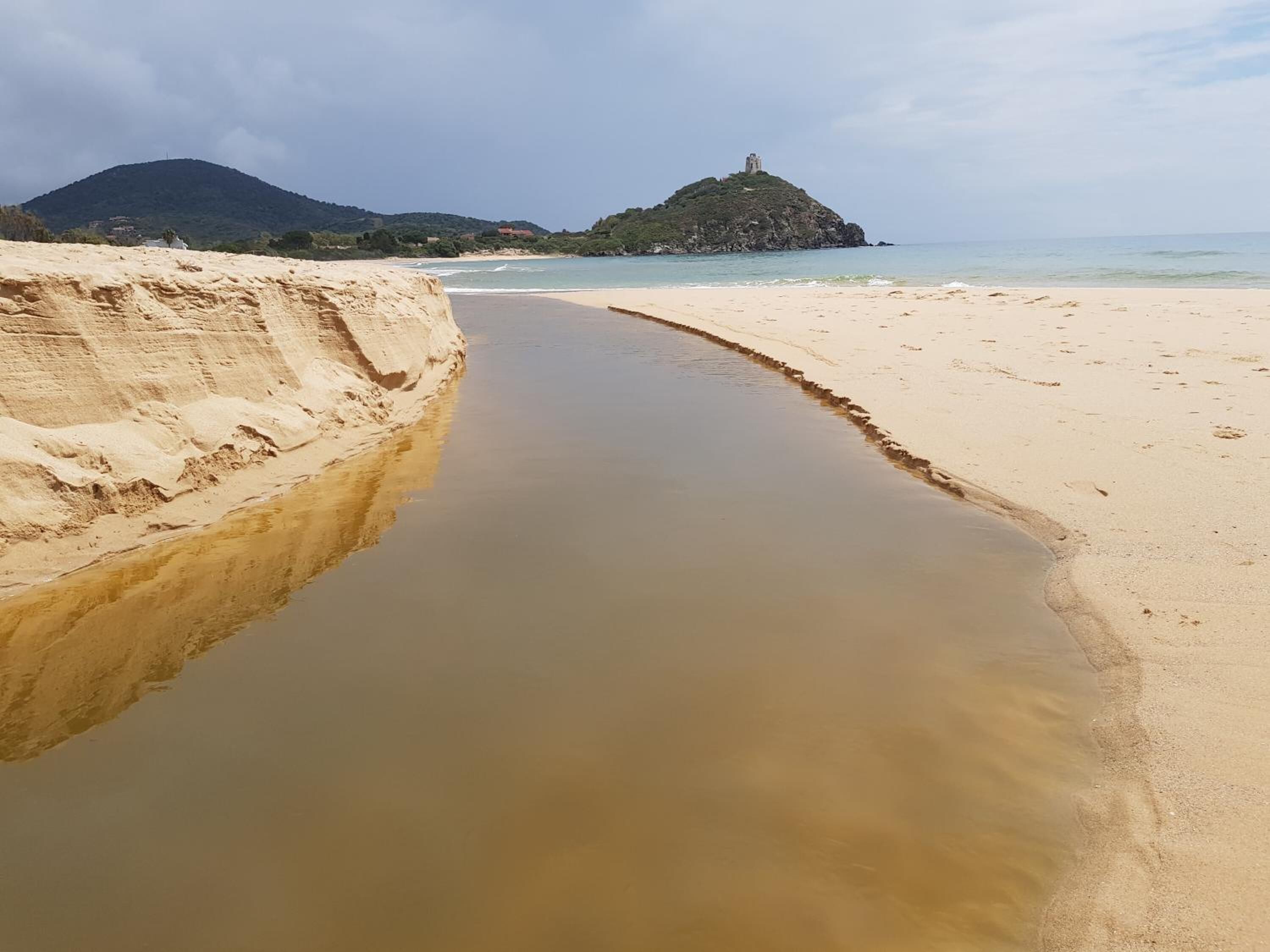 A beach with a water channel, sandy shore, and a hill with a structure in the background.