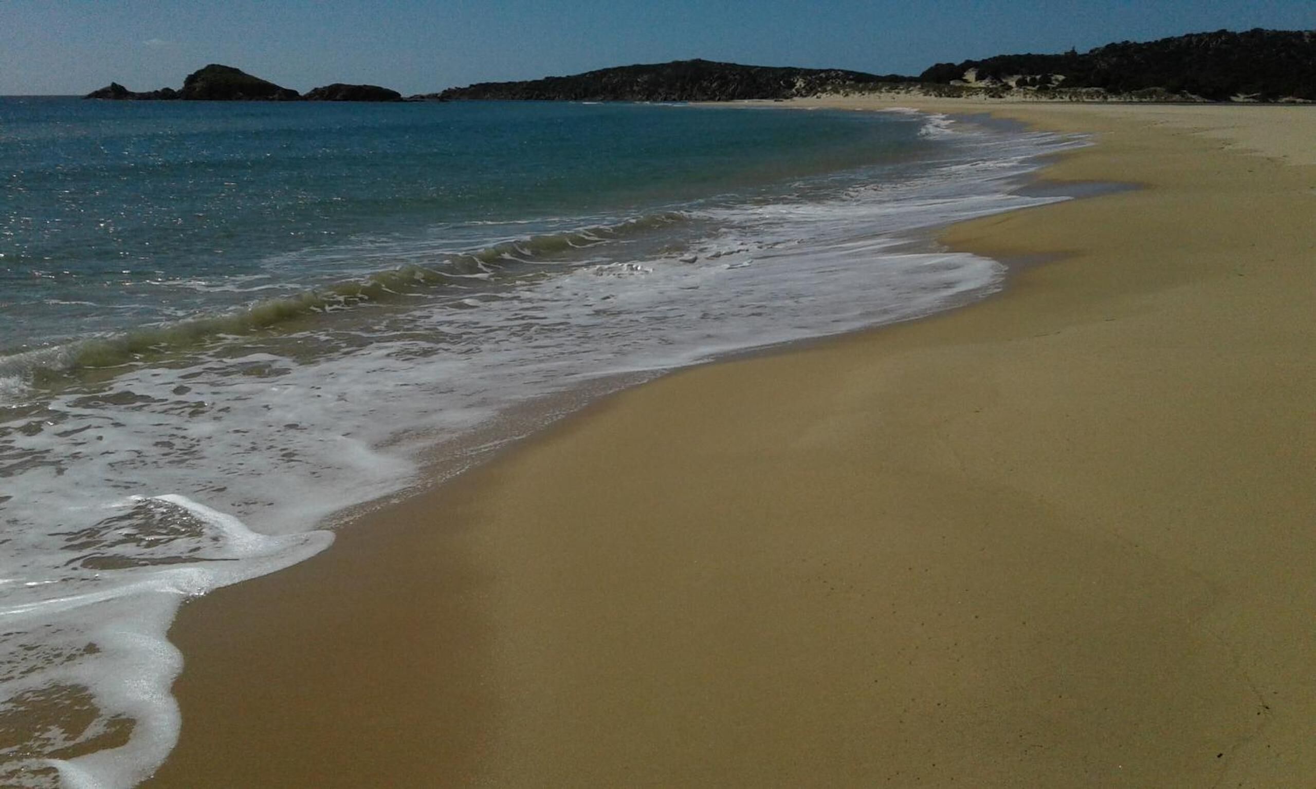 Una playa con agua azul cristalina, orilla de arena y afloramientos rocosos a lo lejos.