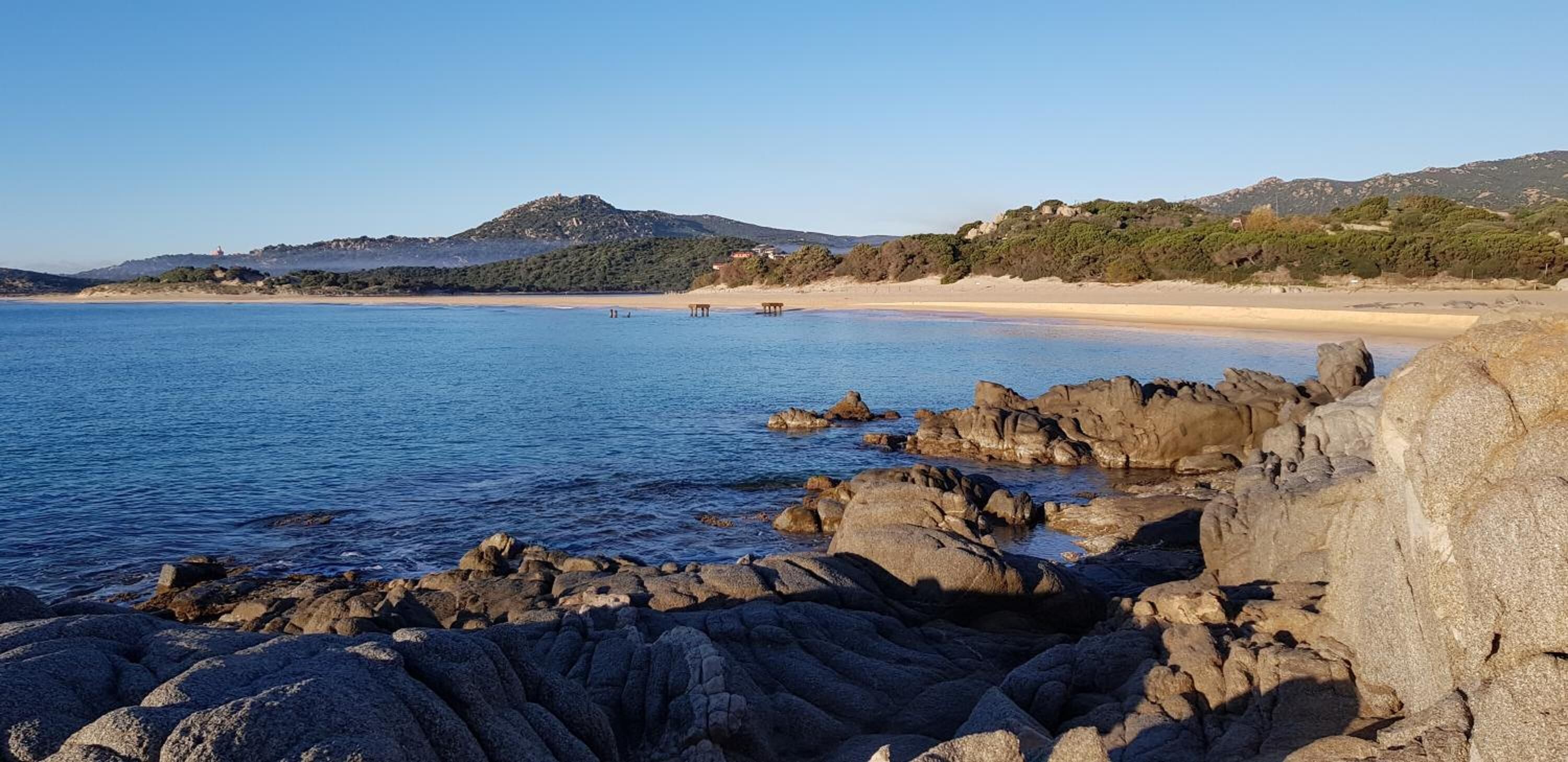 Eine Küstenlandschaft mit felsigen Ufern, einem Sandstrand und Hügeln im Hintergrund.