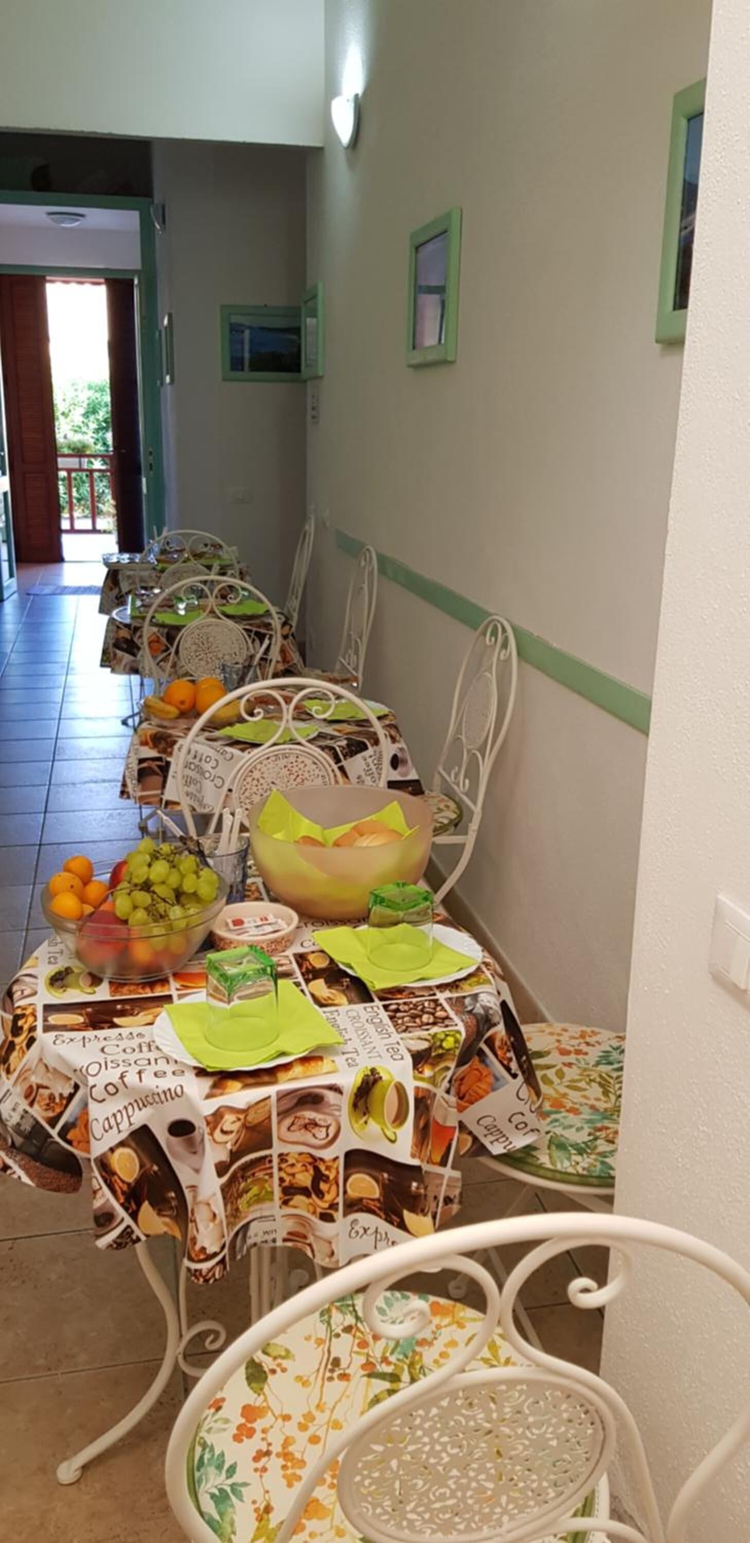 Un comedor con una larga mesa cubierta por un mantel decorado con motivos de café, rodeada de sillas de metal blanco.