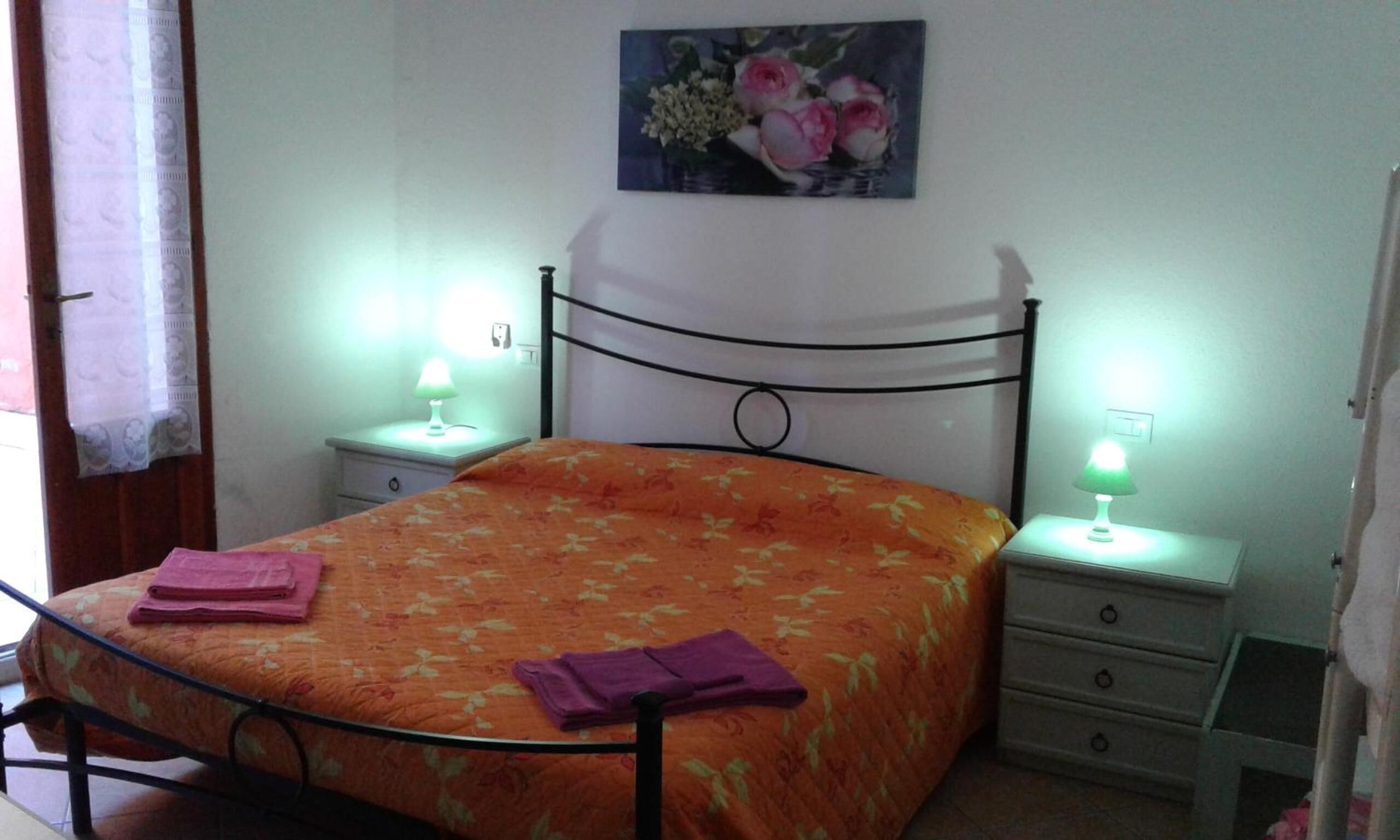 Un dormitori amb un llit, dues taules de nit, una petita taula al costat del llit i un quadre penjat a la paret, i una finestra amb cortines.
