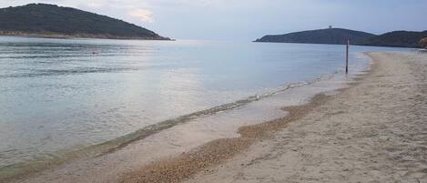 A beach with calm waters, pebbly shore, and distant hills.