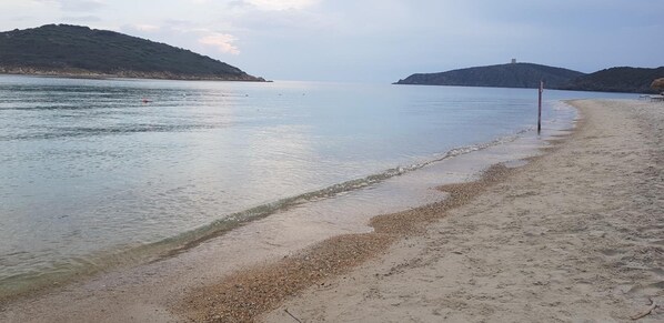 Una playa con aguas tranquilas, orilla pedregosa y colinas a lo lejos.