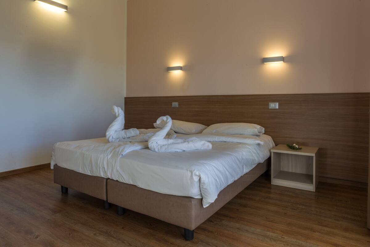 A neatly made bed with white linens and towels arranged in a swan shape, a wooden headboard, and two wall-mounted light fixtures.