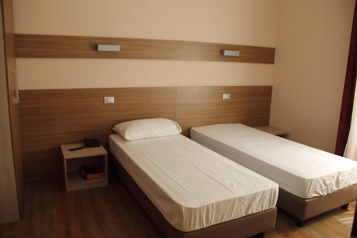 A room with two single beds, a wooden headboard, a nightstand with a lamp, and a small shelf.