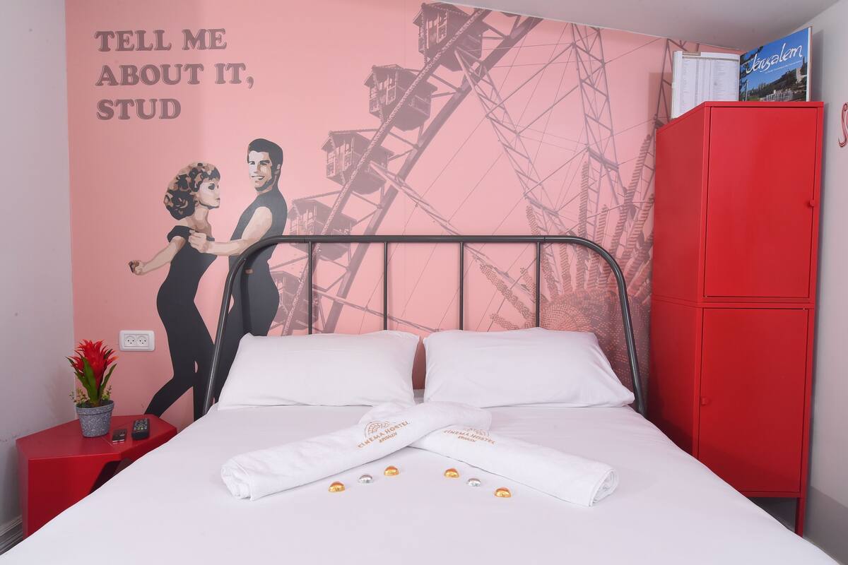 A hotel room with a bed, a red wardrobe, a wall mural, and a small table with a plant and remote controls.