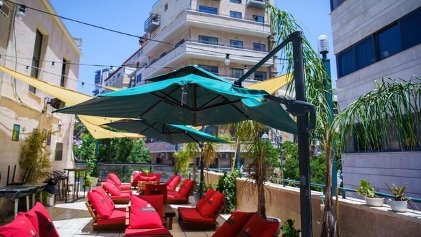 An outdoor seating area with red cushioned chairs and tables, shaded by large umbrellas, set against a backdrop of multi-story buildings. - Cinema Hostel Jerusalem (Jerusalem)