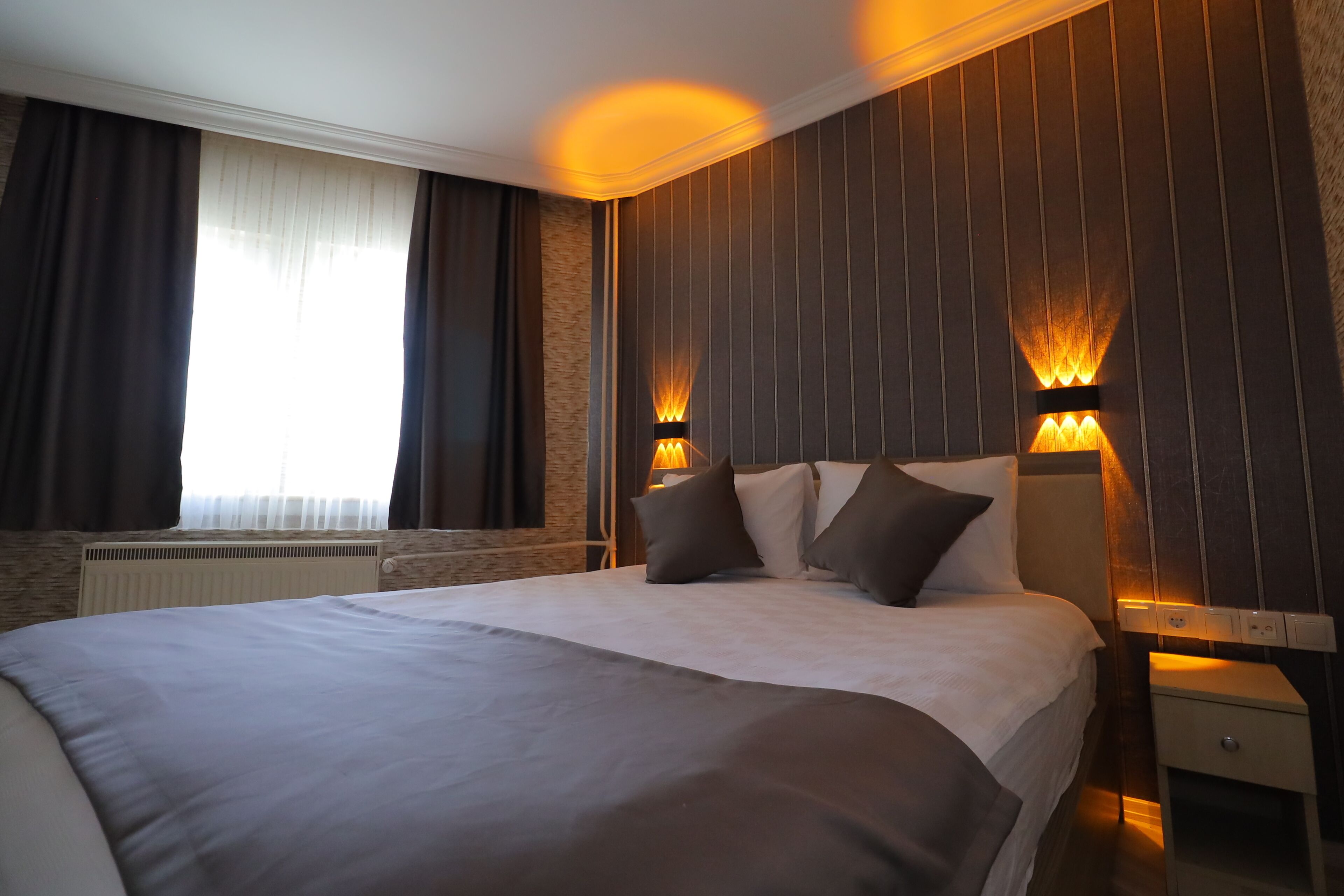 A modern hotel room with a large bed, dark curtains, a radiator, and wall-mounted lighting.