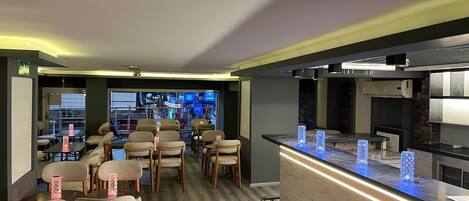 A modern bar area with seating, a counter with illuminated blue glassware, and a view of a cityscape through large windows.
