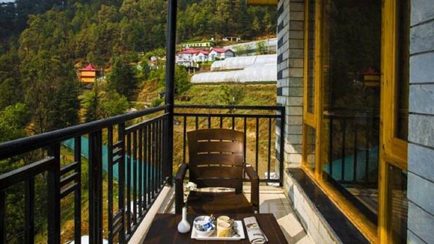 A balcony with a wooden chair, a table with a tray of items, and a view of a hillside with buildings. - Mountain View Resort (Kandaghat)