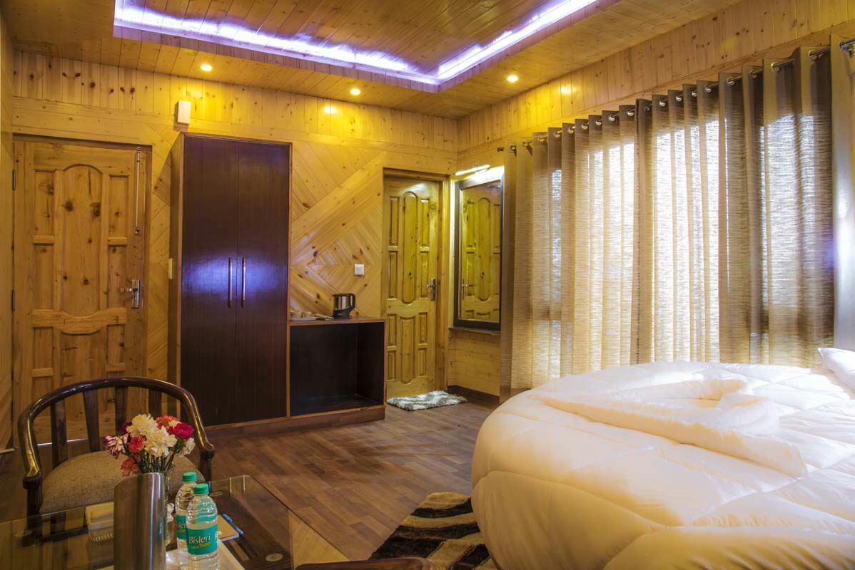 A wooden room with a bed, a desk with a chair, a small table with a vase of flowers, and a door.
