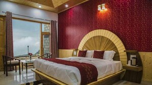 A bedroom with a large bed, a wooden headboard, a nightstand, a table with a chair, and a view through a window. - Mountain View Resort (Kandaghat)