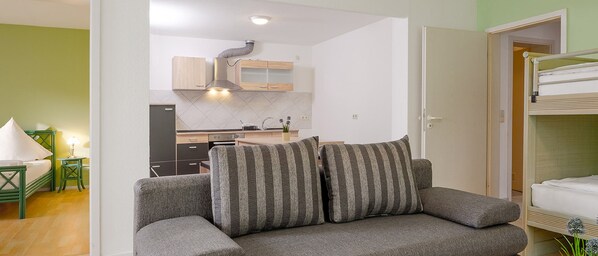 A modern living room with a grey sofa, a small dining area with a kitchen in the background, and a bunk bed in the corner. - Gästehaus Mythos (Rust)