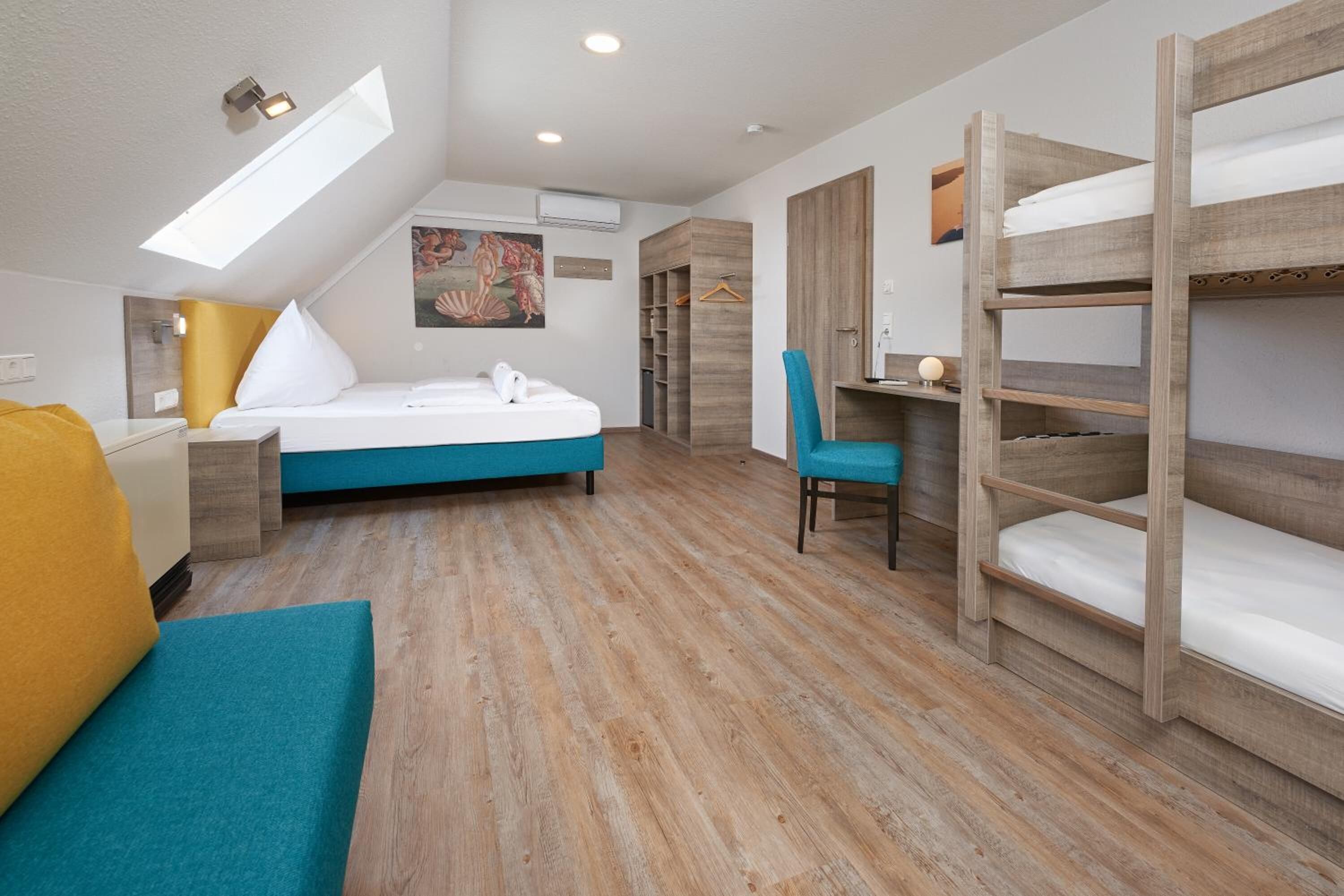 A bedroom with a bunk bed, a desk, a chair, a bed with a blue headboard, a yellow pillow, and a skylight.