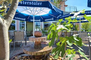 An outdoor seating area with umbrellas labeled "Peterstaler," chairs, a stone fountain, and a building entrance in the background. - Gästehaus Mythos (Rust)