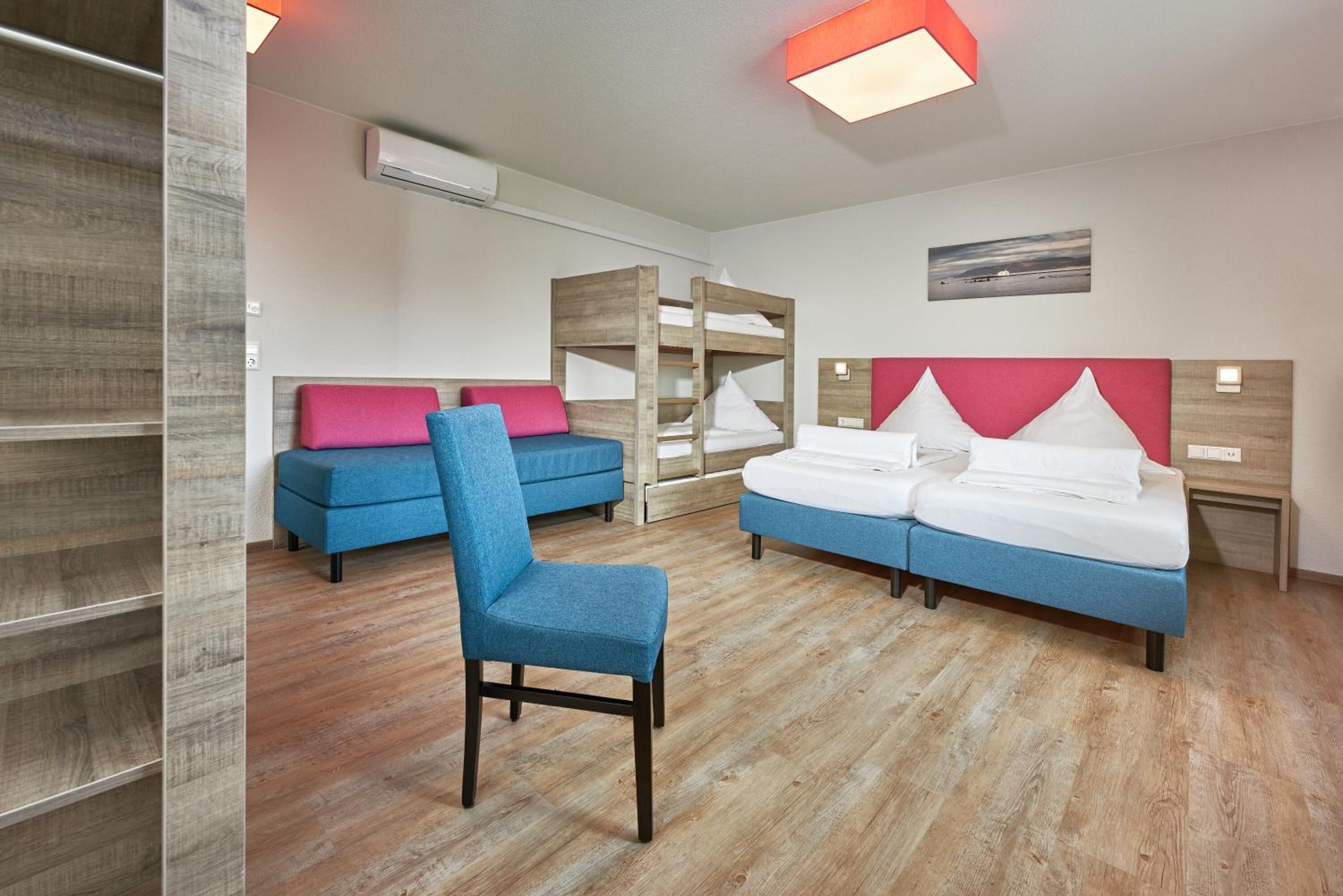 Ein modernes Hotelzimmer mit einem Etagenbett, einem Einzelbett, einem blauen Stuhl und einem Holzschrank.