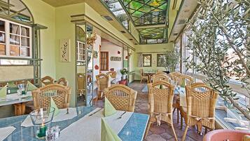 A restaurant with wicker furniture, a glass table, and a view of olive trees.