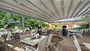 An outdoor patio area with tables and chairs, a covered seating area, and a decorative fire pit. - Gästehaus Mythos (Rust)
