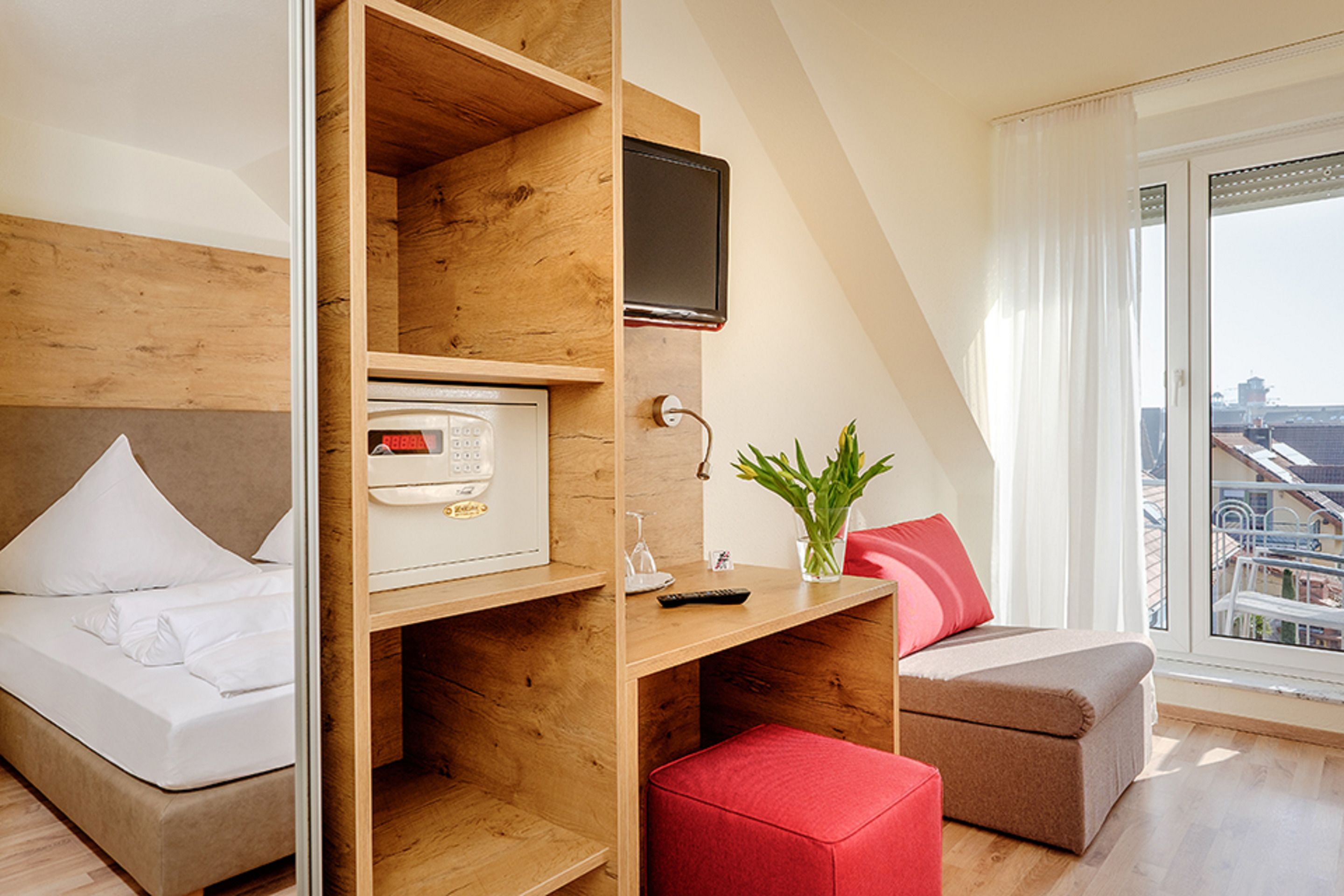 Habitación de hotel con cama, escritorio con televisor, un reposapiés rojo y vista a la ciudad desde la ventana.