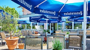 Un área de estar al aire libre con mesas y sillas, cubierta por sombrillas azules con la marca 'Peterstaler', y plantas en macetas.