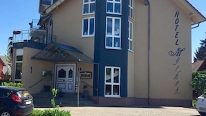 Un hotel de dos plantas con fachada color beige y detalles azules, que destaca por una entrada con escaleras y un pequeño balcón.