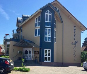 Ett hotell i två våningar med en beige fasad och blå detaljer, med en framträdande entré som inkluderar en trappa och en liten balkong.