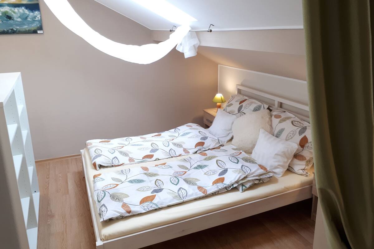 A neatly made bed with a leaf-patterned bedspread, a bedside lamp, and a painting on the wall.