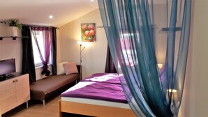 A bedroom with a bed, bedside table, TV, and a window with curtains. - Hédi Vendégház (Eger)