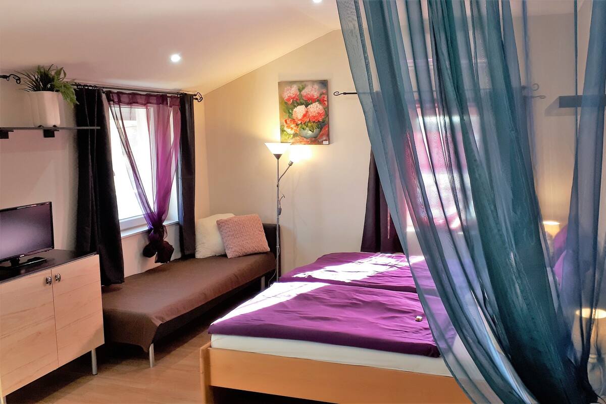 A bedroom with a bed, bedside table, TV, and a window with curtains.