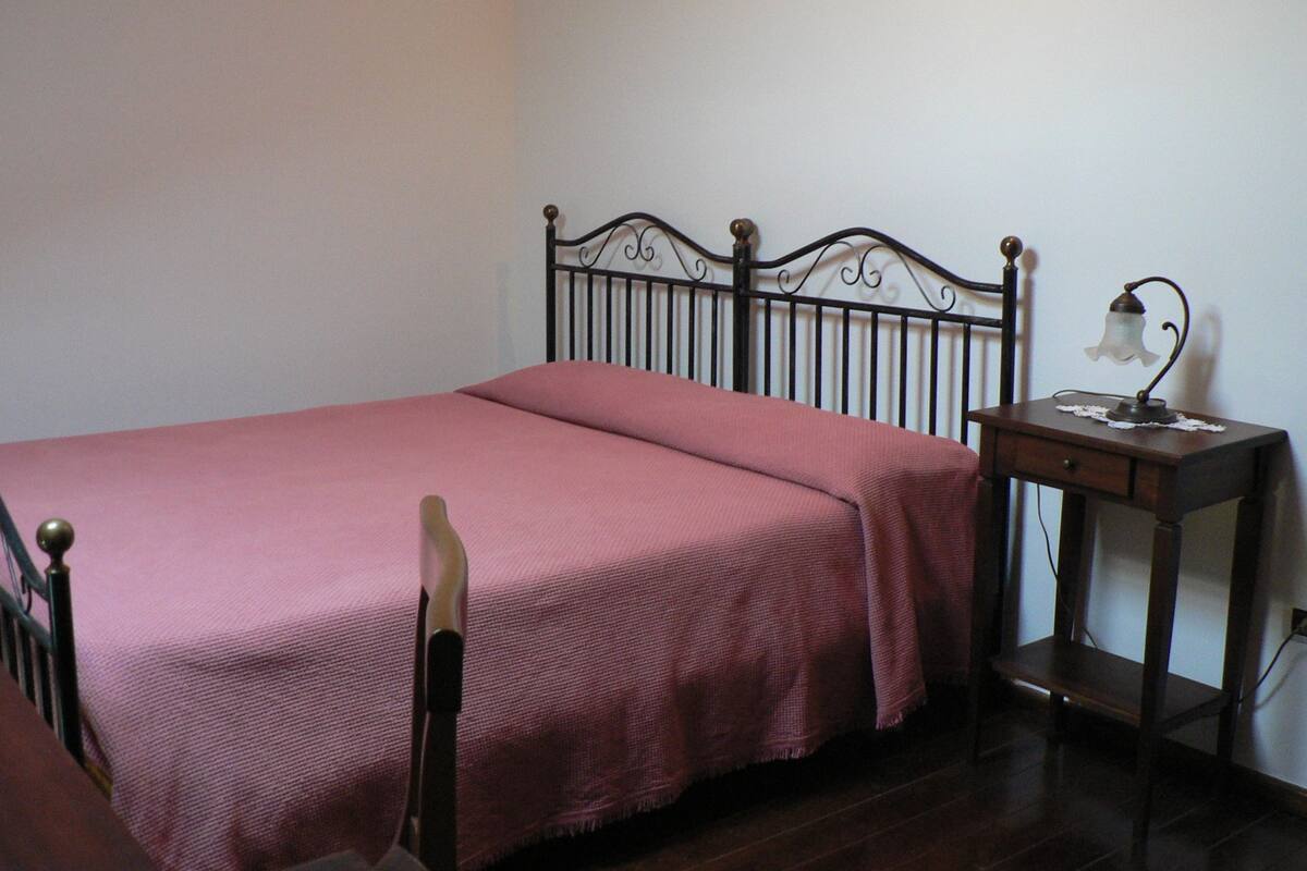 Una cama individual con cabecera de metal, una mesita de noche con lámpara y una pared lisa.