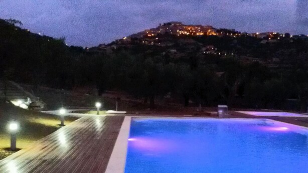 Una piscina con cubierta de madera e iluminación, rodeada de árboles y una colina con luces al fondo.
