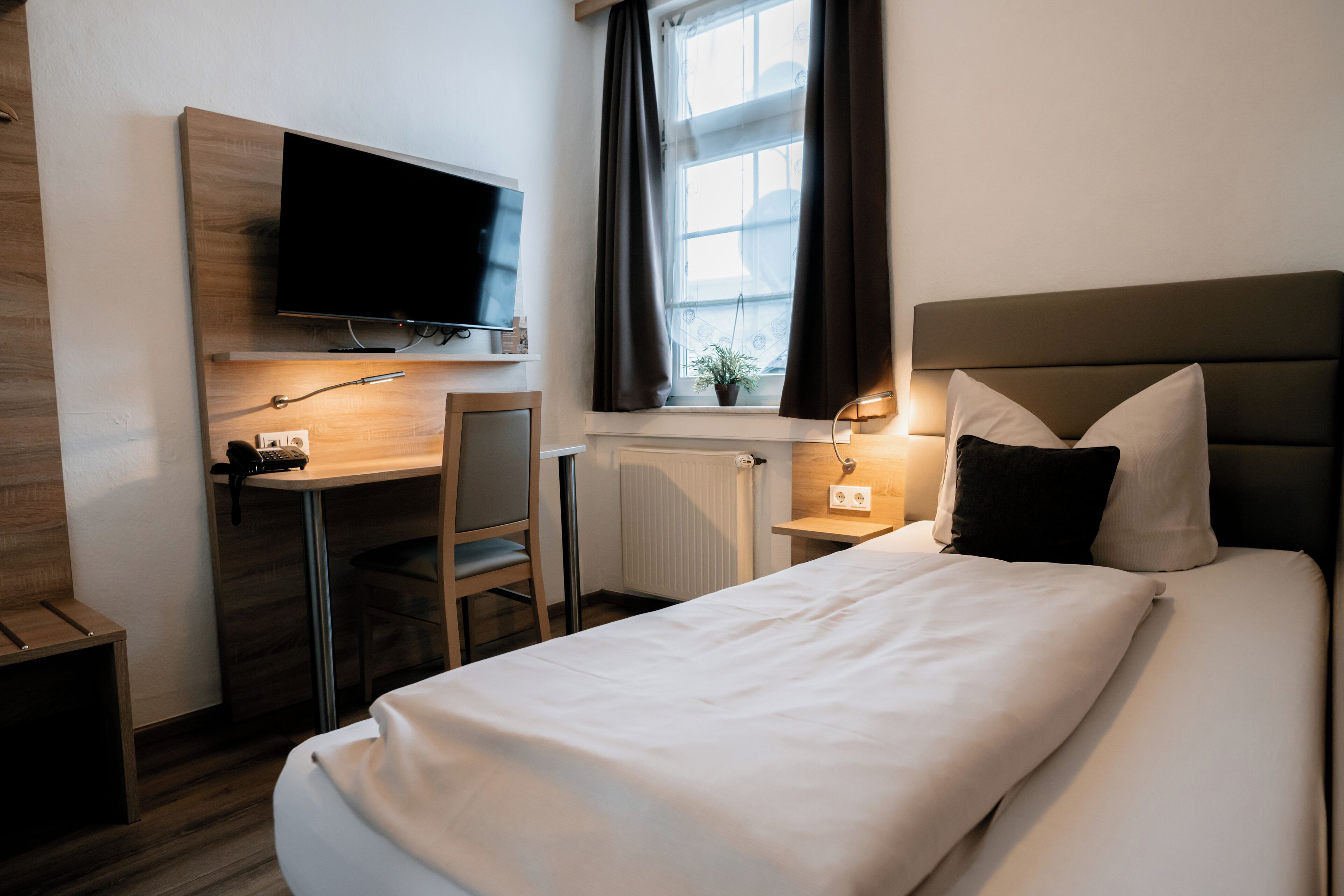 A hotel room with a bed, a desk with a chair, a television mounted on the wall, and a window with curtains.