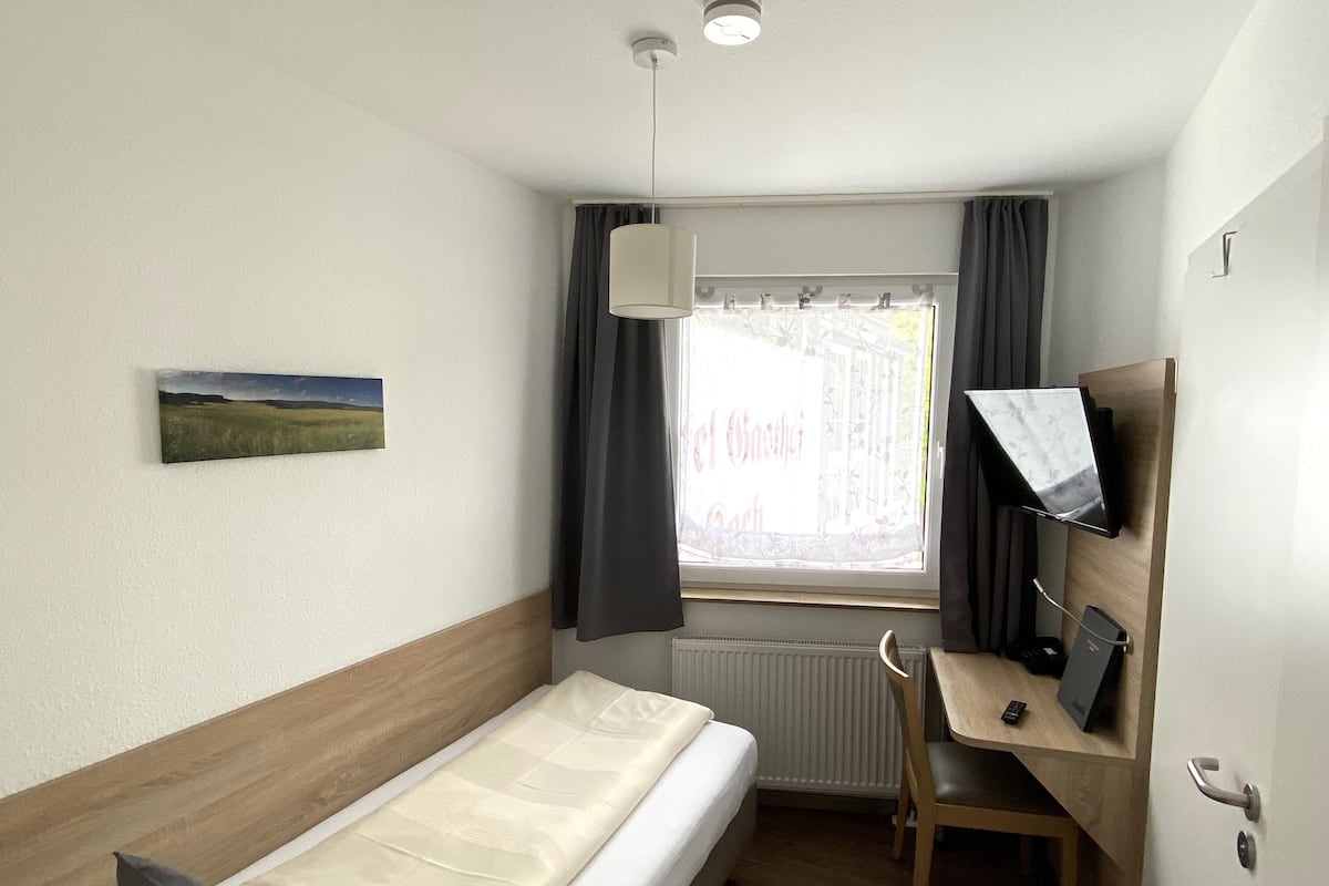 A compact hotel room with a single bed, a desk with a chair, a small window with curtains, a radiator, and a mounted TV.