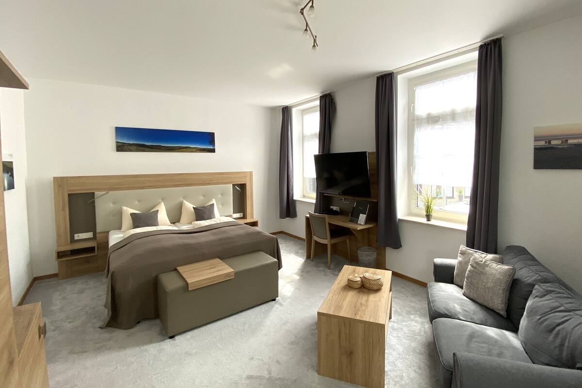 A modern hotel room with a bed, a sofa, a TV, and large windows.
