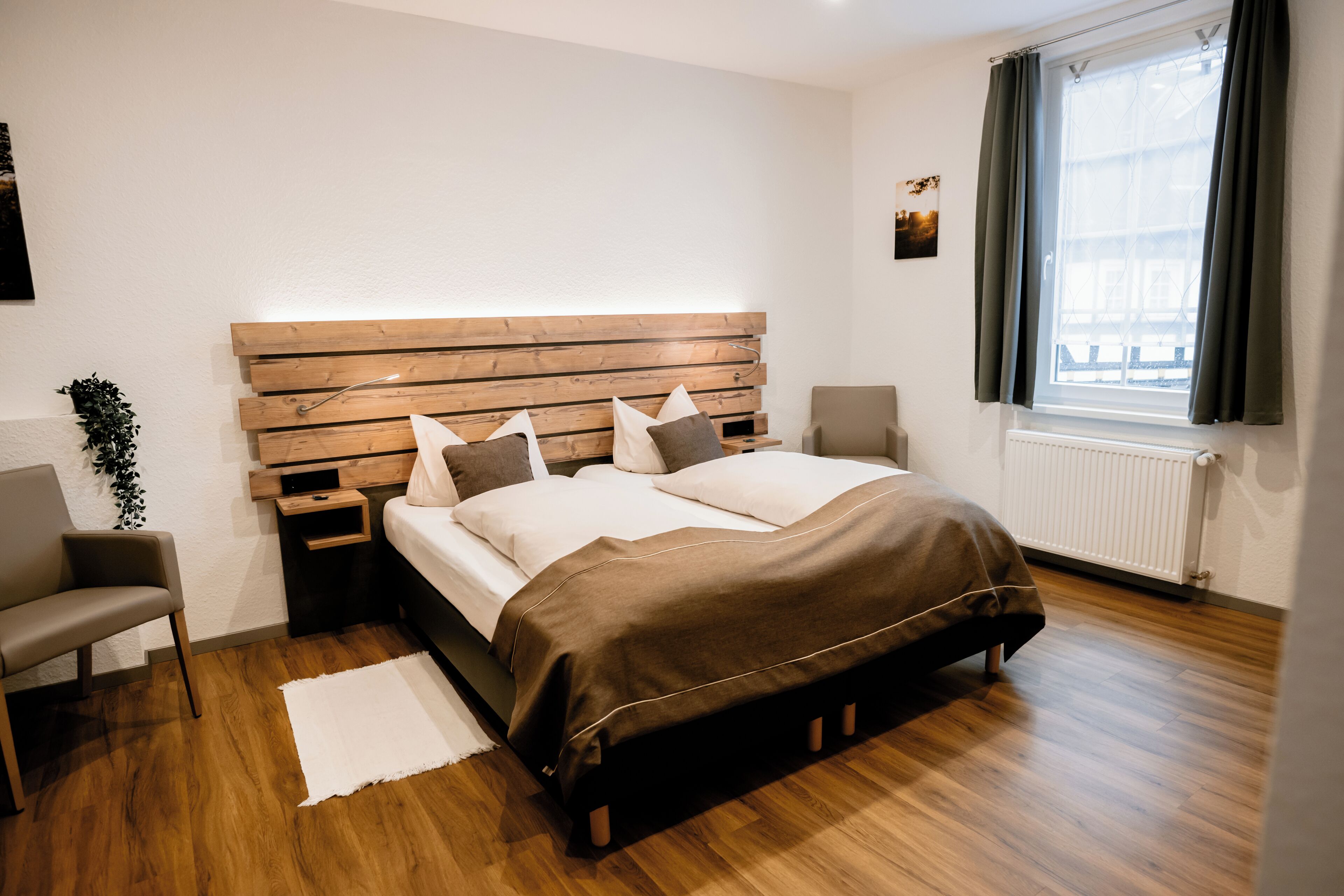 Un dormitorio moderno con cabecera de madera, cama con cubrecama marrón, mesita de noche, una silla y una ventana con cortinas.