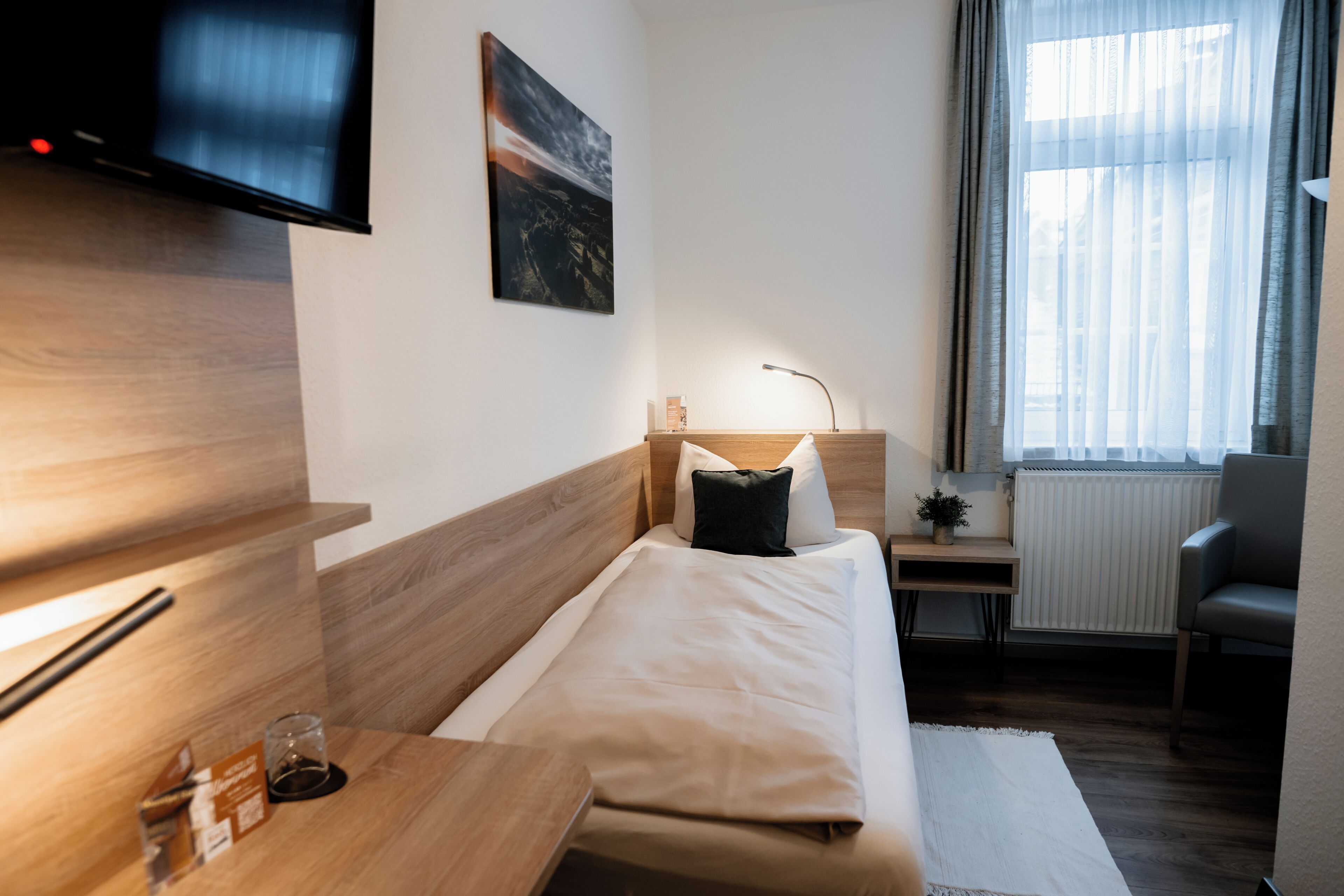 Habitación de hotel con cama, mesita de noche, lámpara, silla y televisor montado en la pared.