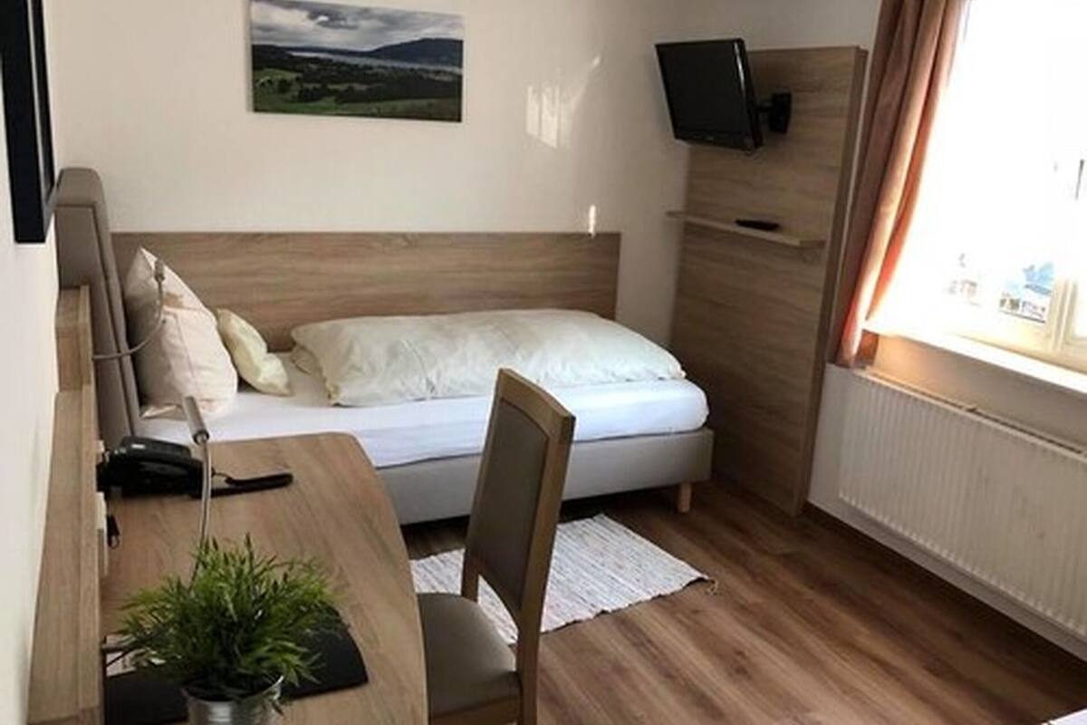 A small, neatly arranged bedroom with a bed, a desk, a chair, a TV mounted on the wall, and a window with curtains.