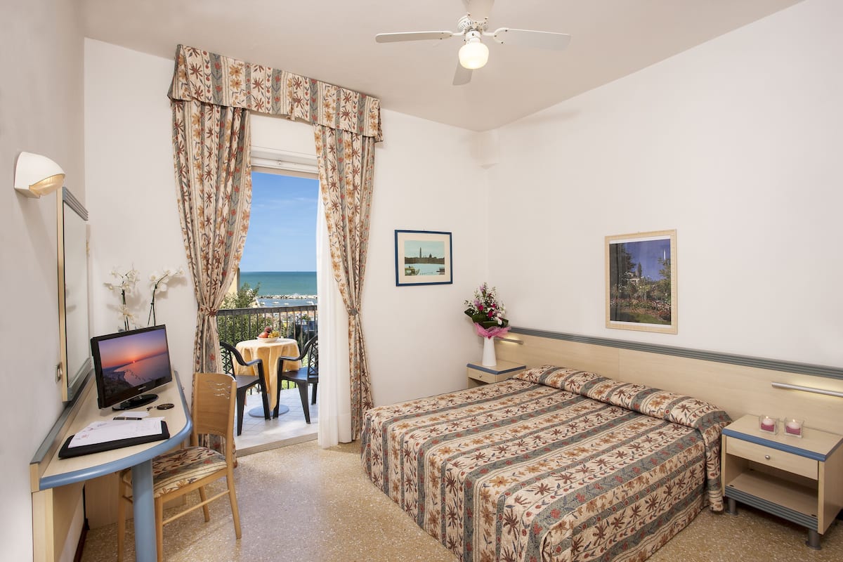 A bedroom with a bed, a desk with a computer, a chair, a nightstand, and a window with curtains.