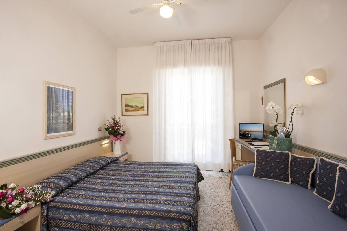 A hotel room with a bed, a sofa, a desk with a computer, and a window with curtains.