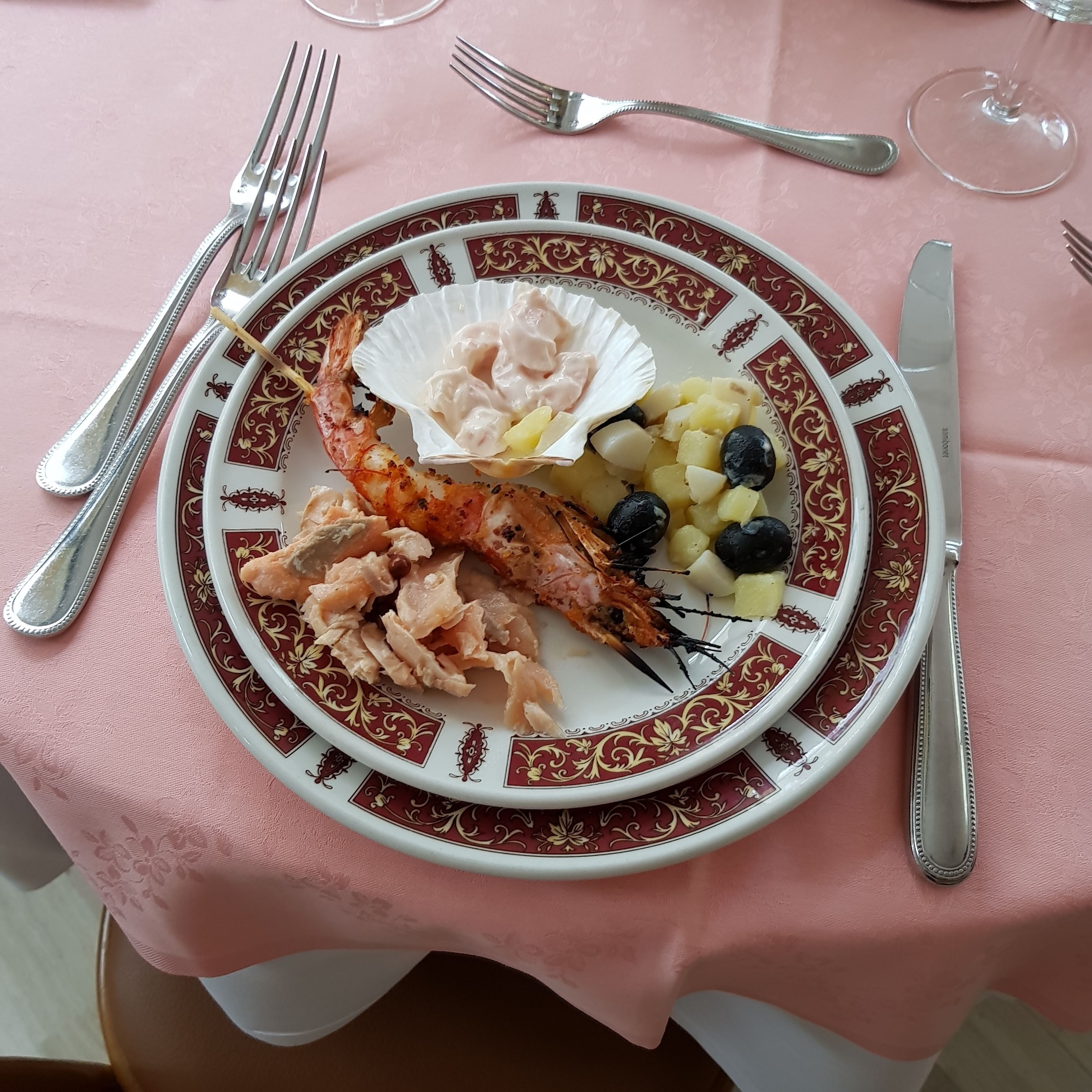 A plate of seafood including shrimp, scallops, and potatoes with olives, served on a decorative plate with a pink tablecloth.
