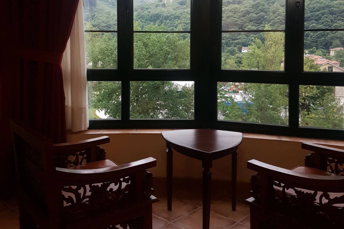 Ein Zimmer mit einem großen Fenster mit Blick auf eine gebirgige Landschaft. Im Zimmer befinden sich zwei Holzstühle und ein kleiner Tisch.