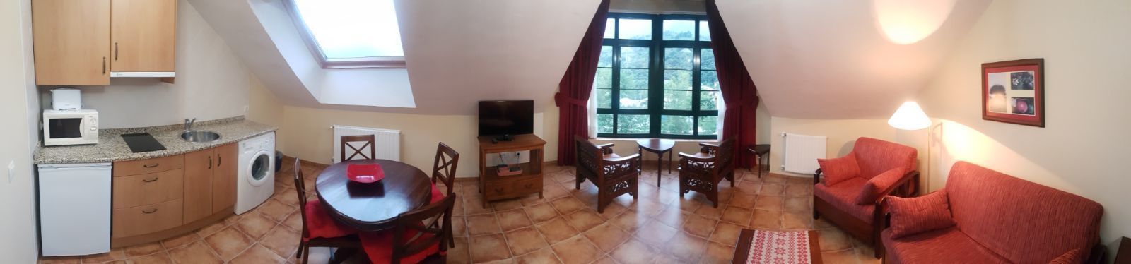 A compact living space with a kitchen, dining area, and seating area. It includes a washing machine, a microwave, a TV, and a large window providing natural light.